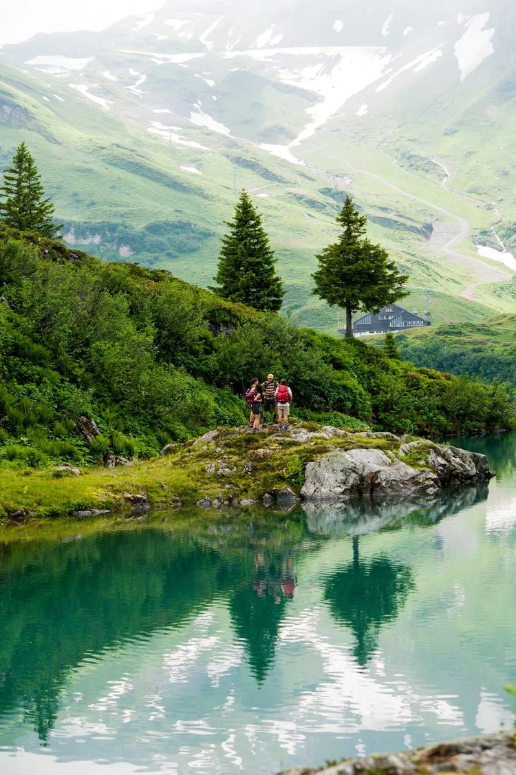 Three hikers with backpacks standing on a small rocky outcrop by a green lake surrounded by lush greenery and pine trees, with snow-capped mountains in the background.