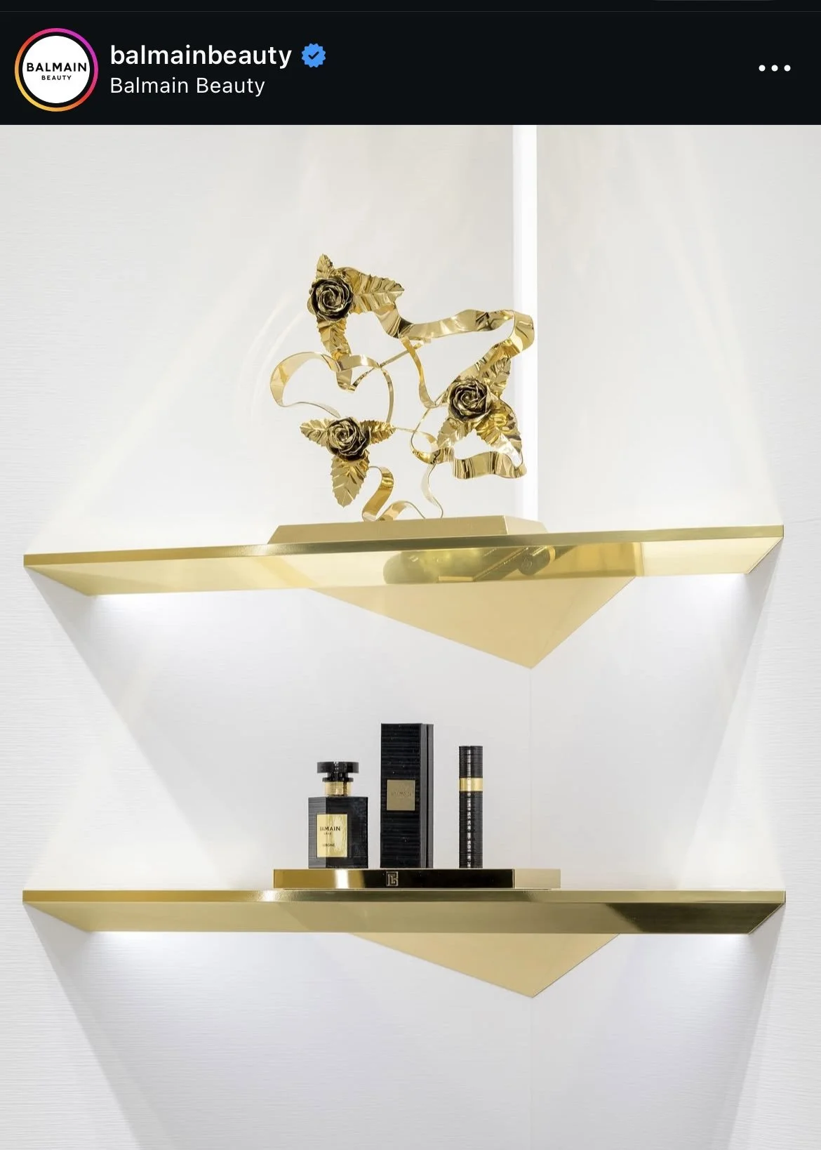 A decorative display on a white wall featuring two gold-colored shelves with gold accents. The top shelf has an abstract gold sculpture with roses and leaves, and the bottom shelf holds black and gold perfume bottles.