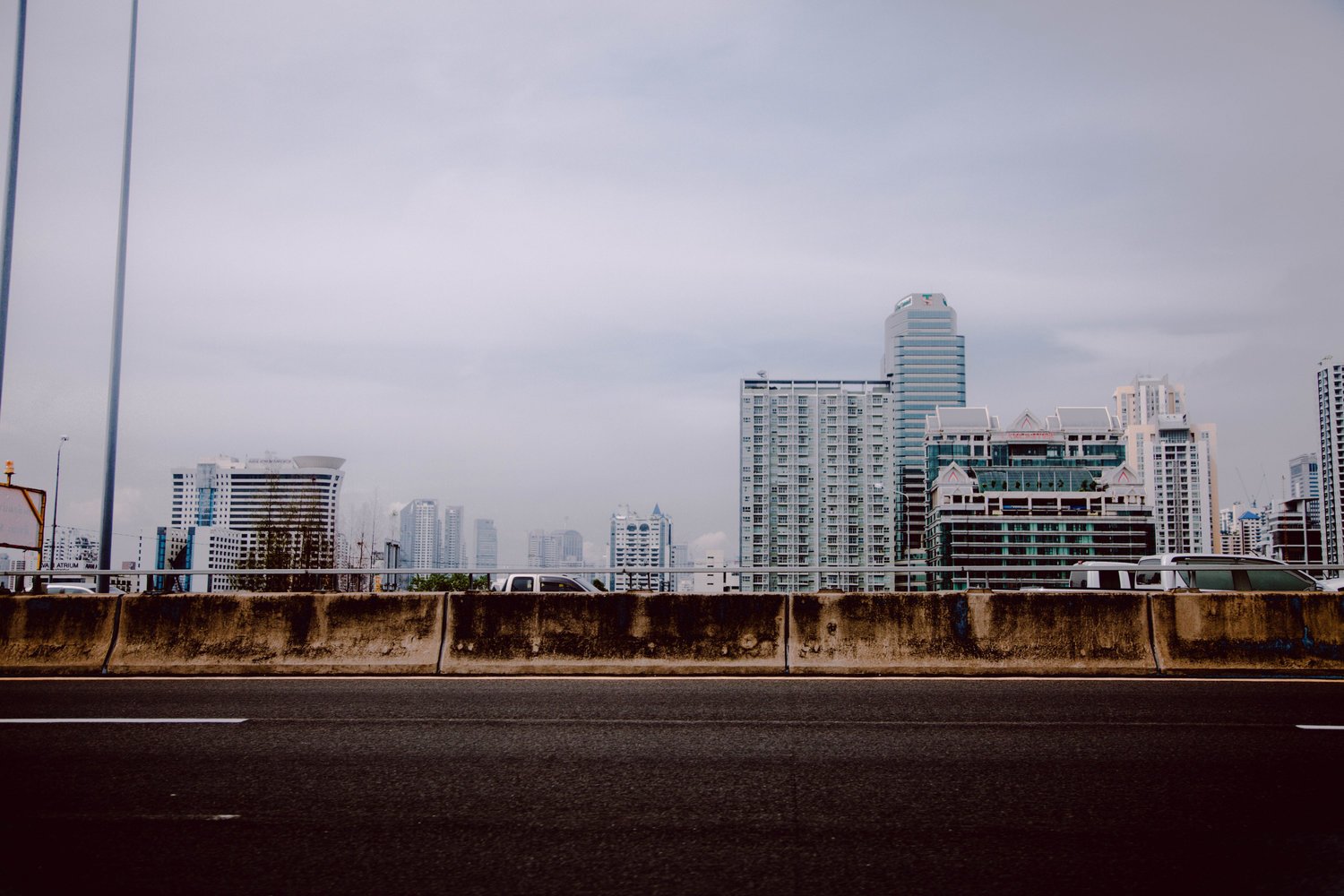 View of a city skyline with skyscrapers behind a concrete highway barrier, under an overcast sky.