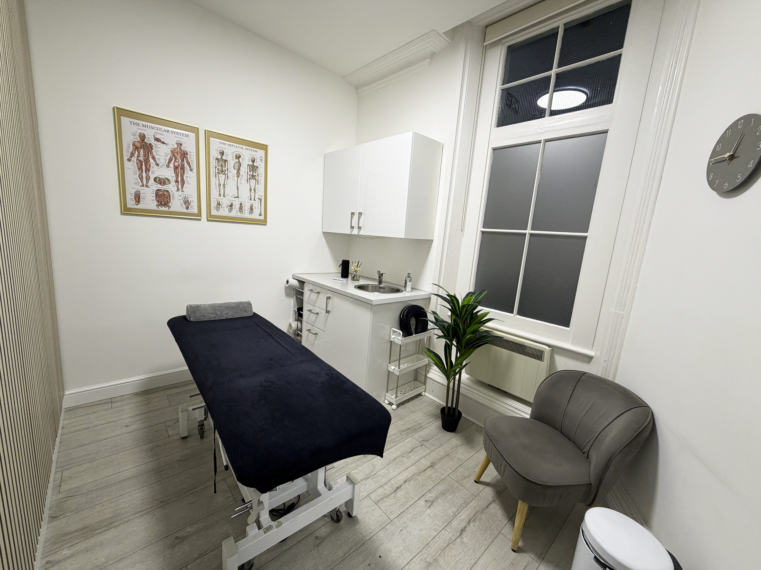 Medical examination room with a massage table, a chair, a large window, and medical posters on the wall.
