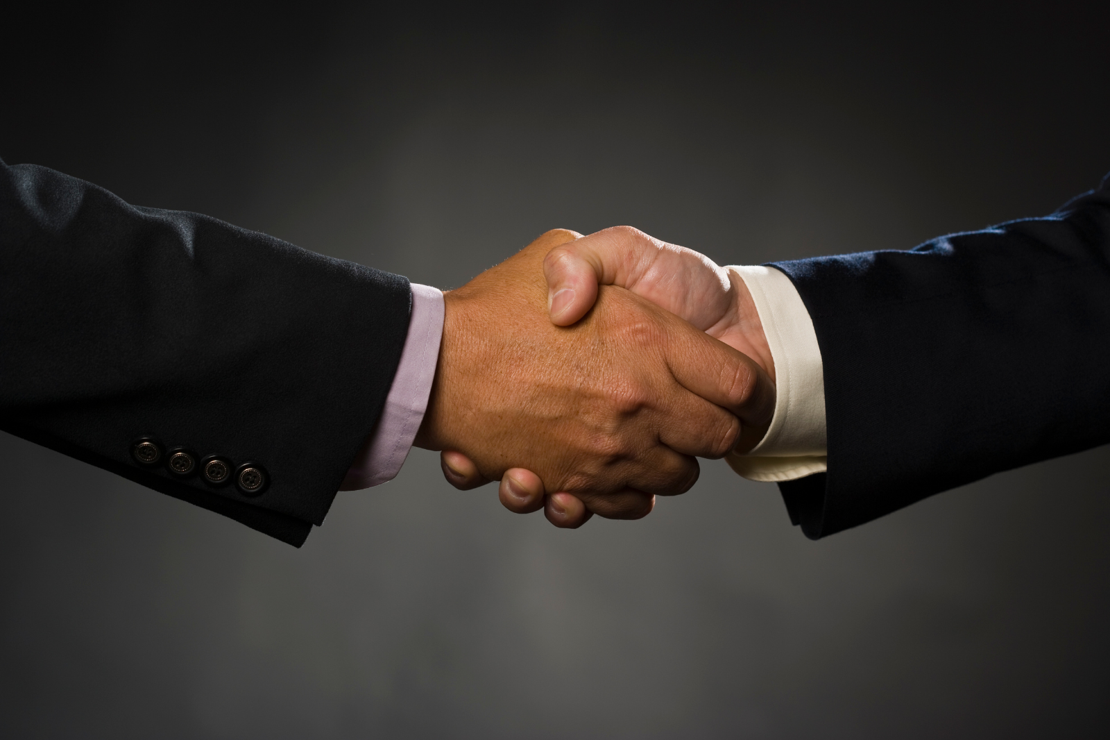 Close-up of two people shaking hands, both wearing dark suits, against a dark background.