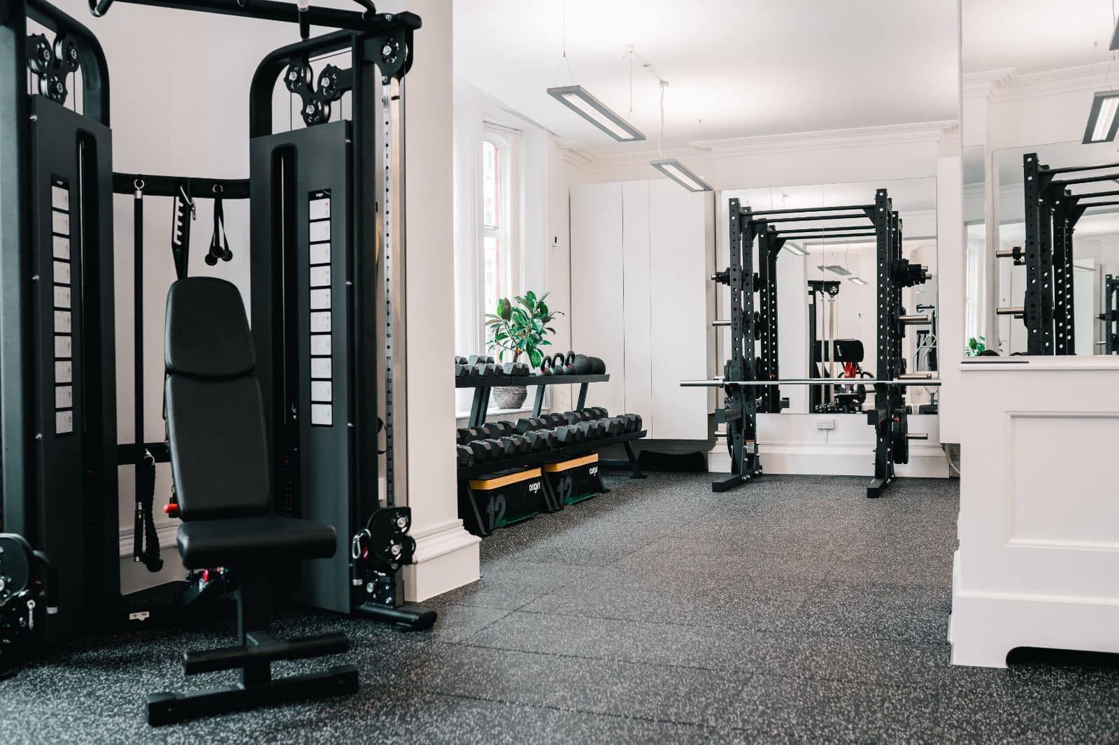 Empty gym with weight lifting equipment, including a multi-station machine, free weights, and a bench press, with large windows and a mirror.