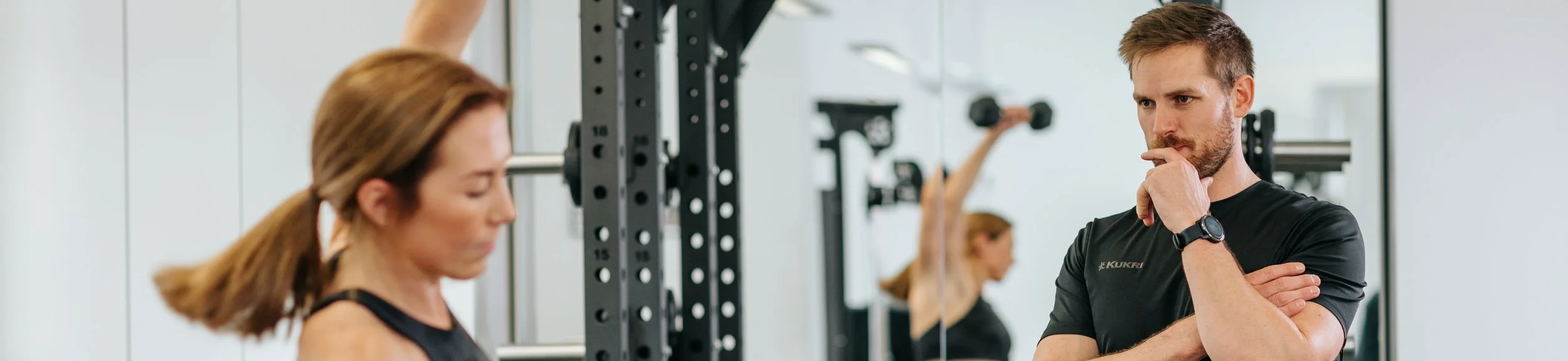 A personal trainer or coach talking to a woman at the gym, with another woman exercising in the background.
