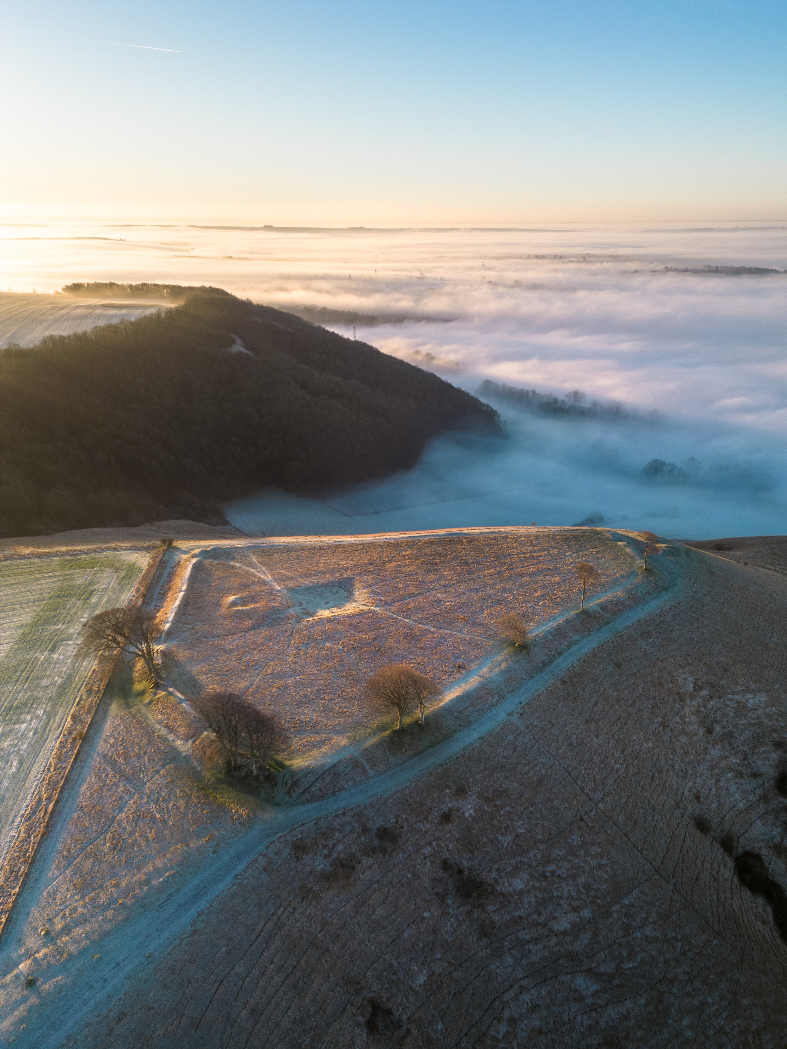Aerial view of a frosty landscape with an ancient hillfort, surrounded by fields and partially covered by fog, during sunrise.