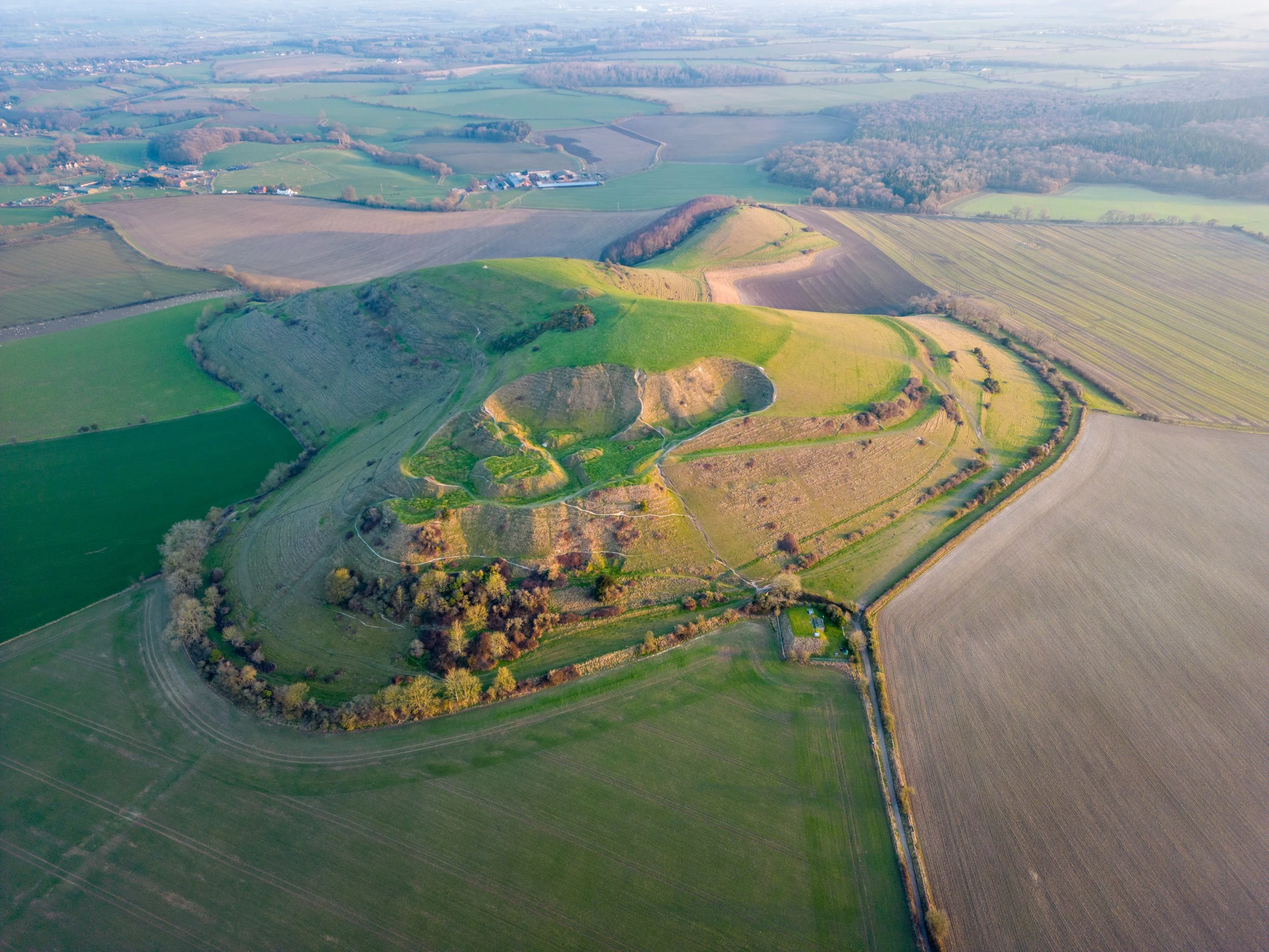 Aerial view of Hambledon Hill in Dorset, England, featuring an ancient hillfort surrounded by green and brown agricultural fields.