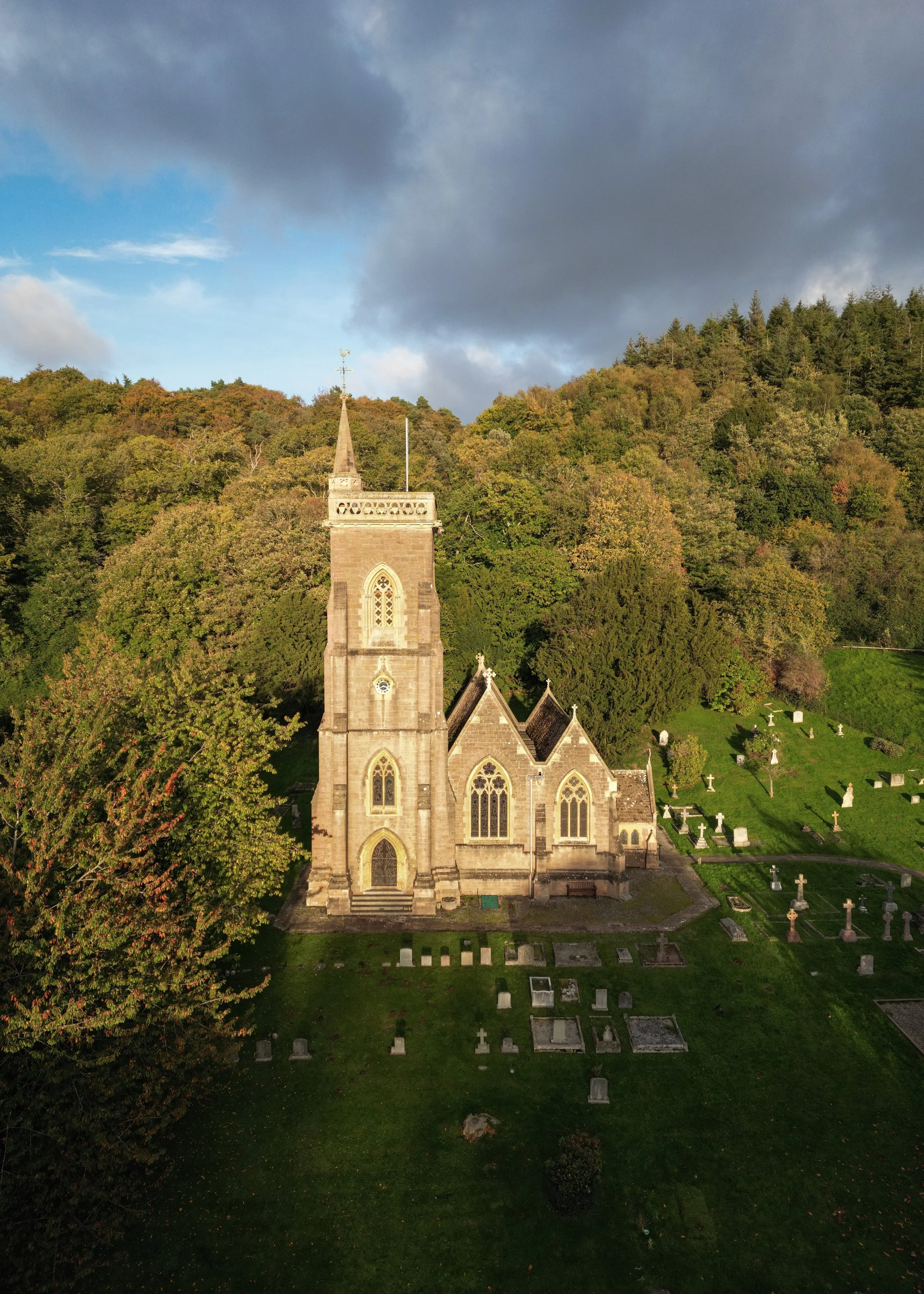 A historic stone church with a tall steeple and pointed arched windows, surrounded by a graveyard with headstones, set against a hillside covered in trees and a sky with dark and light clouds.