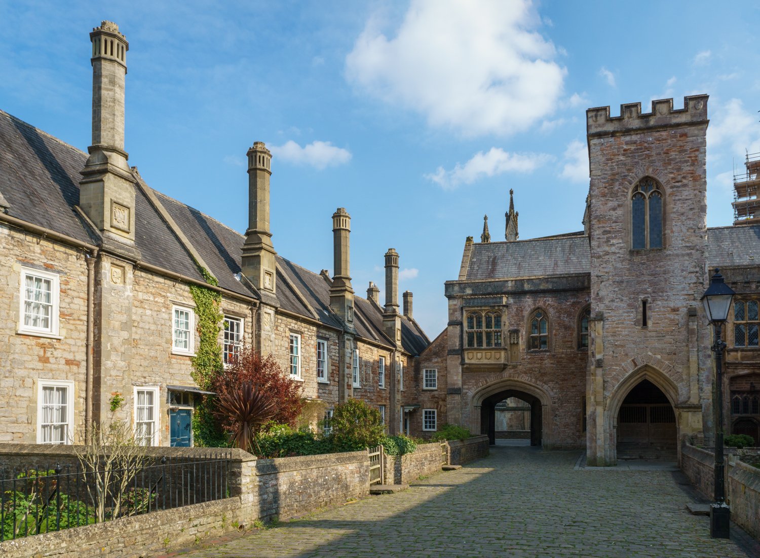 A historic stone building with a gated archway, traditional chimney pots, and a cobblestone street in front, under a blue sky with some clouds.
