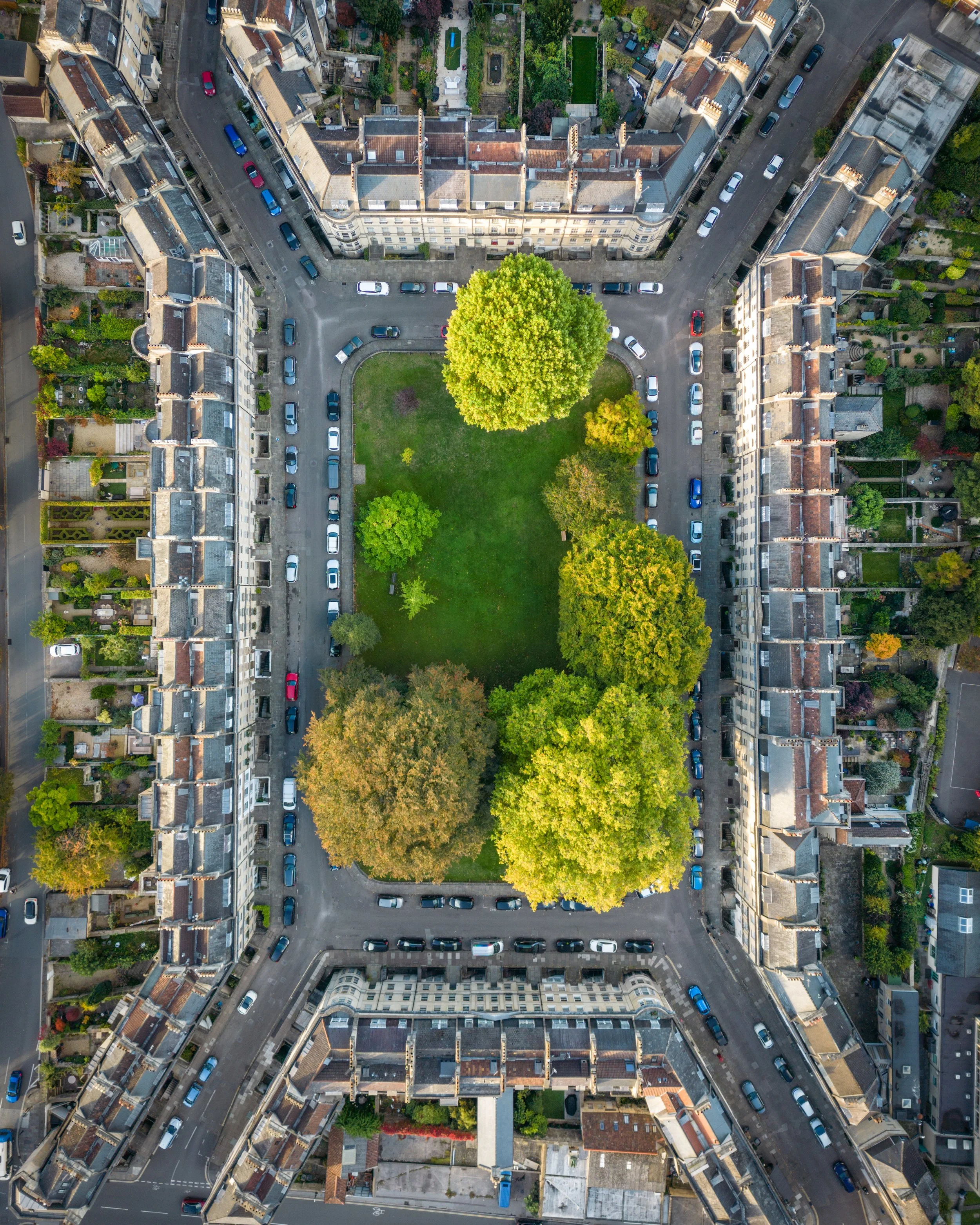 An aerial view of a central courtyard with grass and trees, surrounded by multi-story residential buildings with parking spaces and cars along the streets.