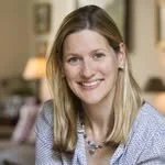 A young woman with long blonde hair wearing a light blue shirt, smiling indoors.