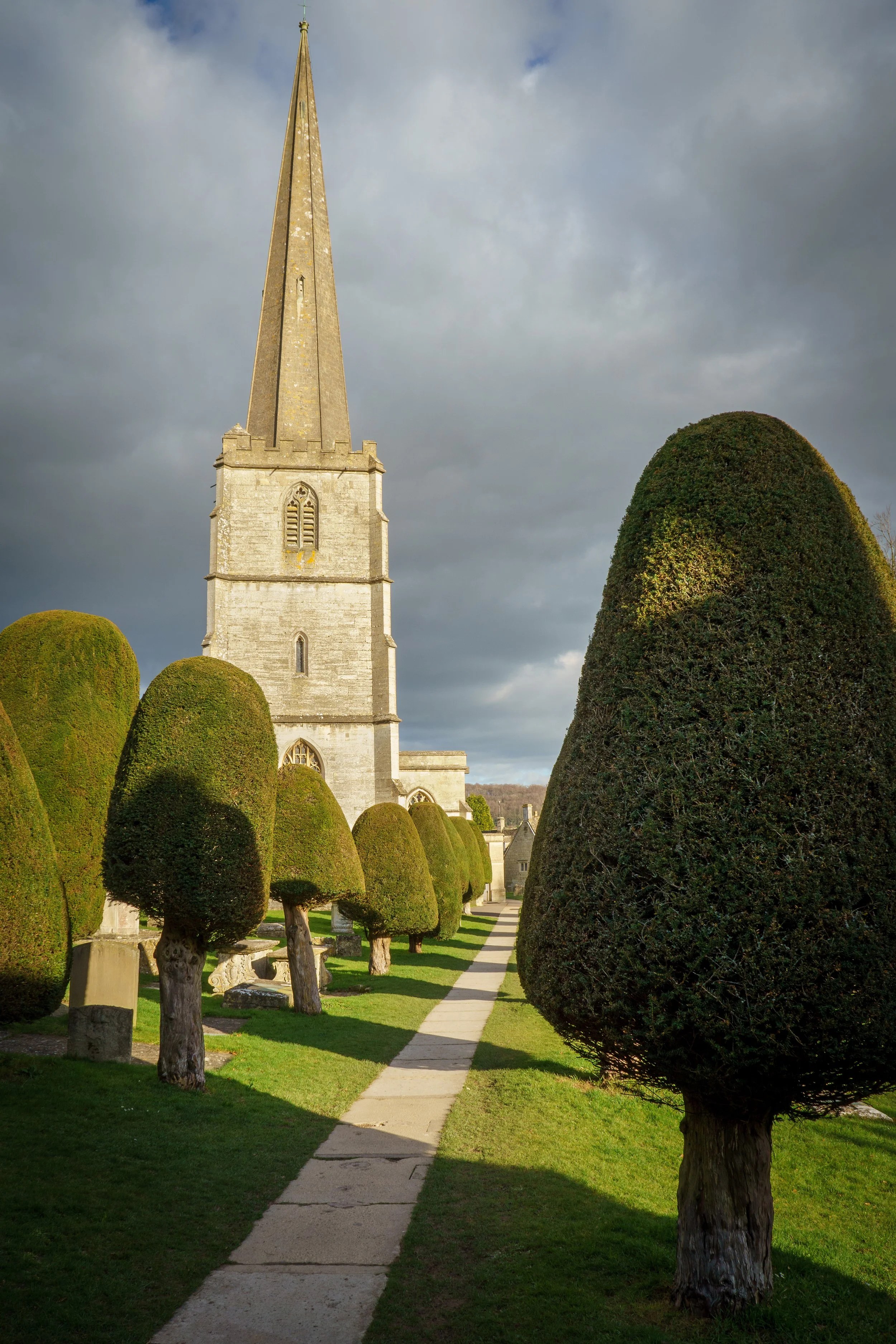 A church with a tall steeple surrounded by manicured bushes and a paved walkway, under a cloudy sky.