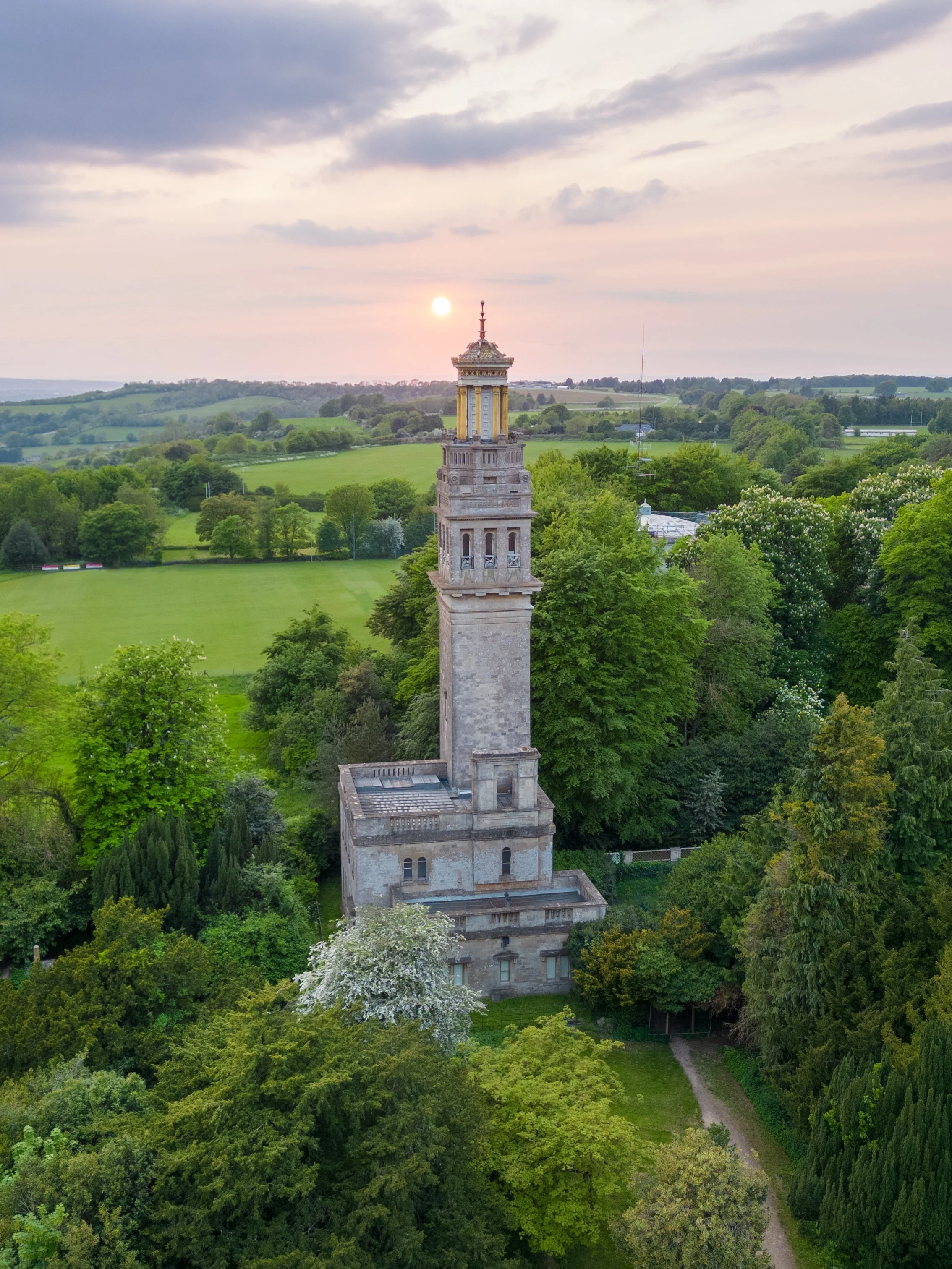 A tall, historic stone tower with a decorative top stands among lush green trees in a rural landscape at sunset, with rolling hills and a partly cloudy sky in the background.