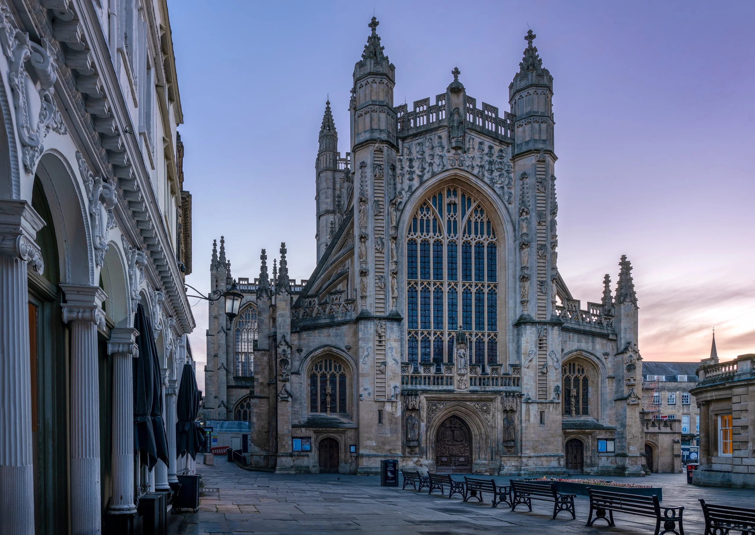 A Gothic-style cathedral with large stained glass windows, decorative stone carvings, and tall towers, situated in a quiet town square with benches and buildings around.