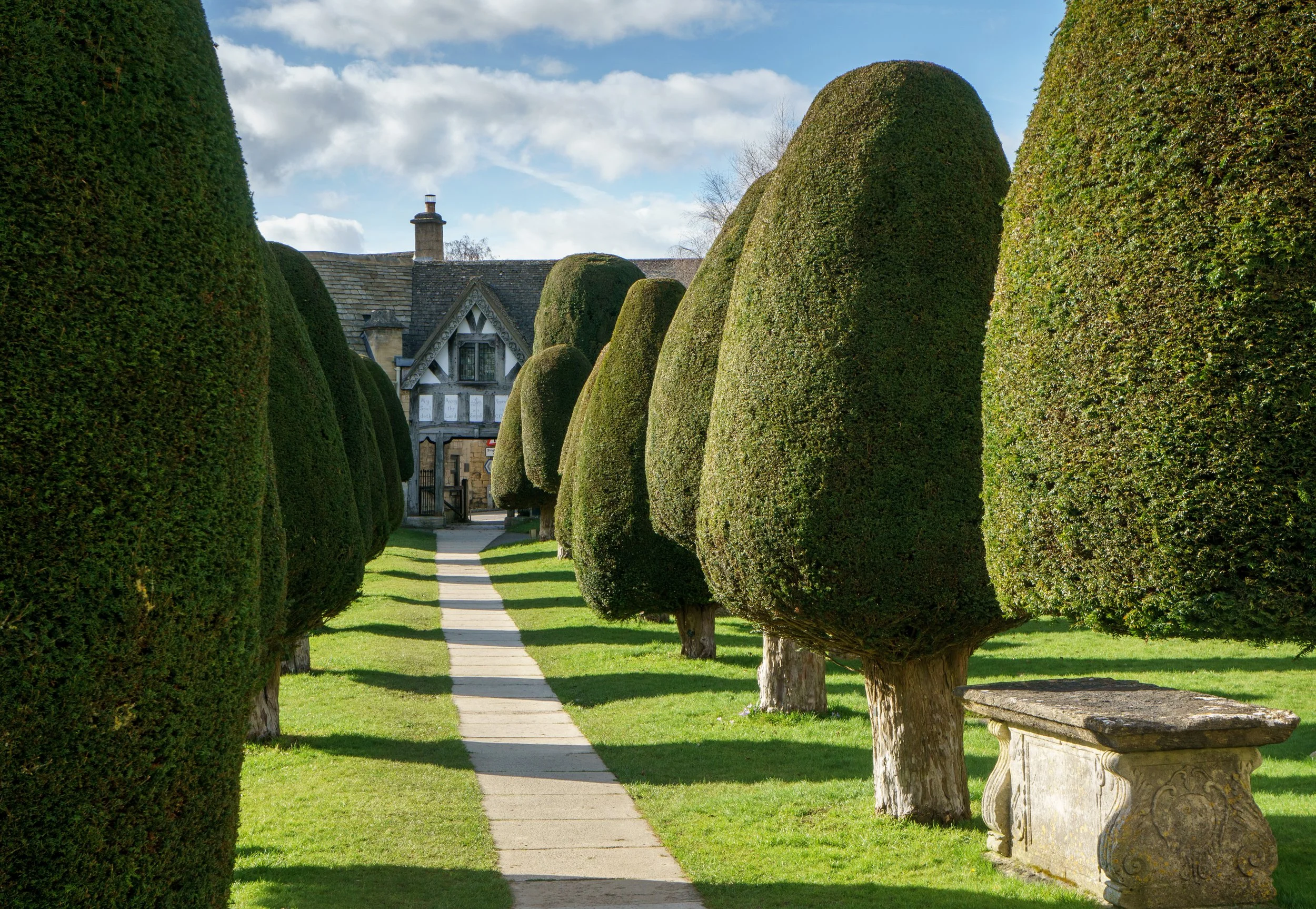 A pathway lined with neatly trimmed, tall, rounded green bushes leading to a traditional house with a chimney and stone exterior, under a partly cloudy sky.