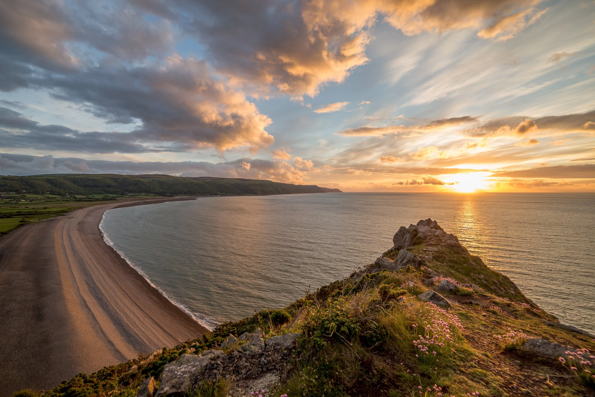 Scenic coastal view with rocky coastline, beach, and sunset over the ocean, framed by clouds and grassy hills.