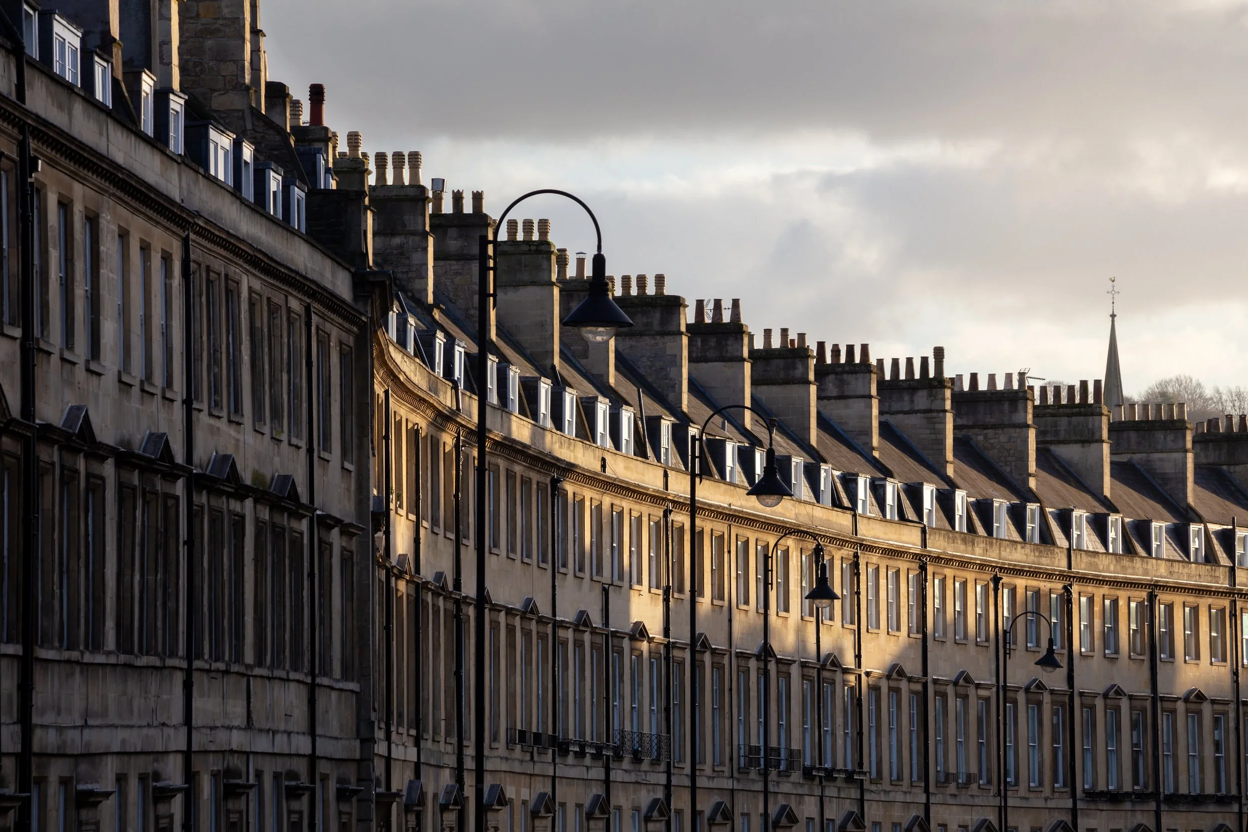 Row of historic European buildings with chimney stacks and street lamps, illuminated by soft sunlight, under a cloudy sky.