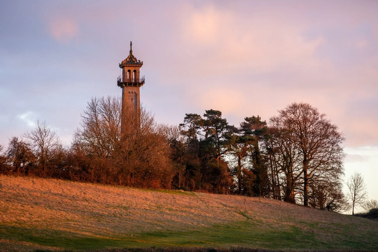 A tall stone tower with decorative top, surrounded by trees, on a grassy hill at sunset.