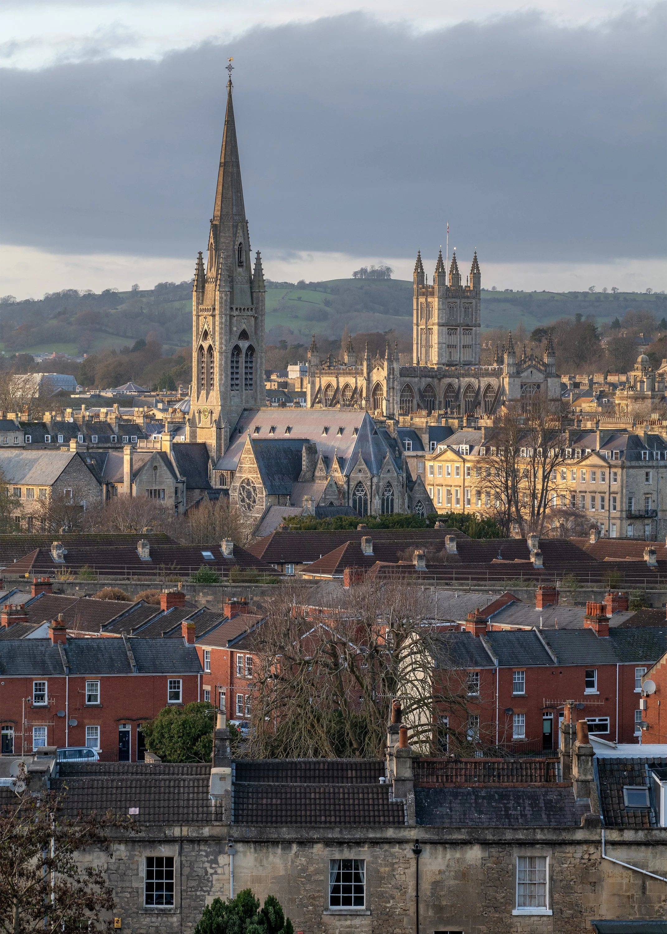 View of a historic city with a large Gothic church with a tall spire and many smaller buildings. Rolling hills are in the background under a cloudy sky.