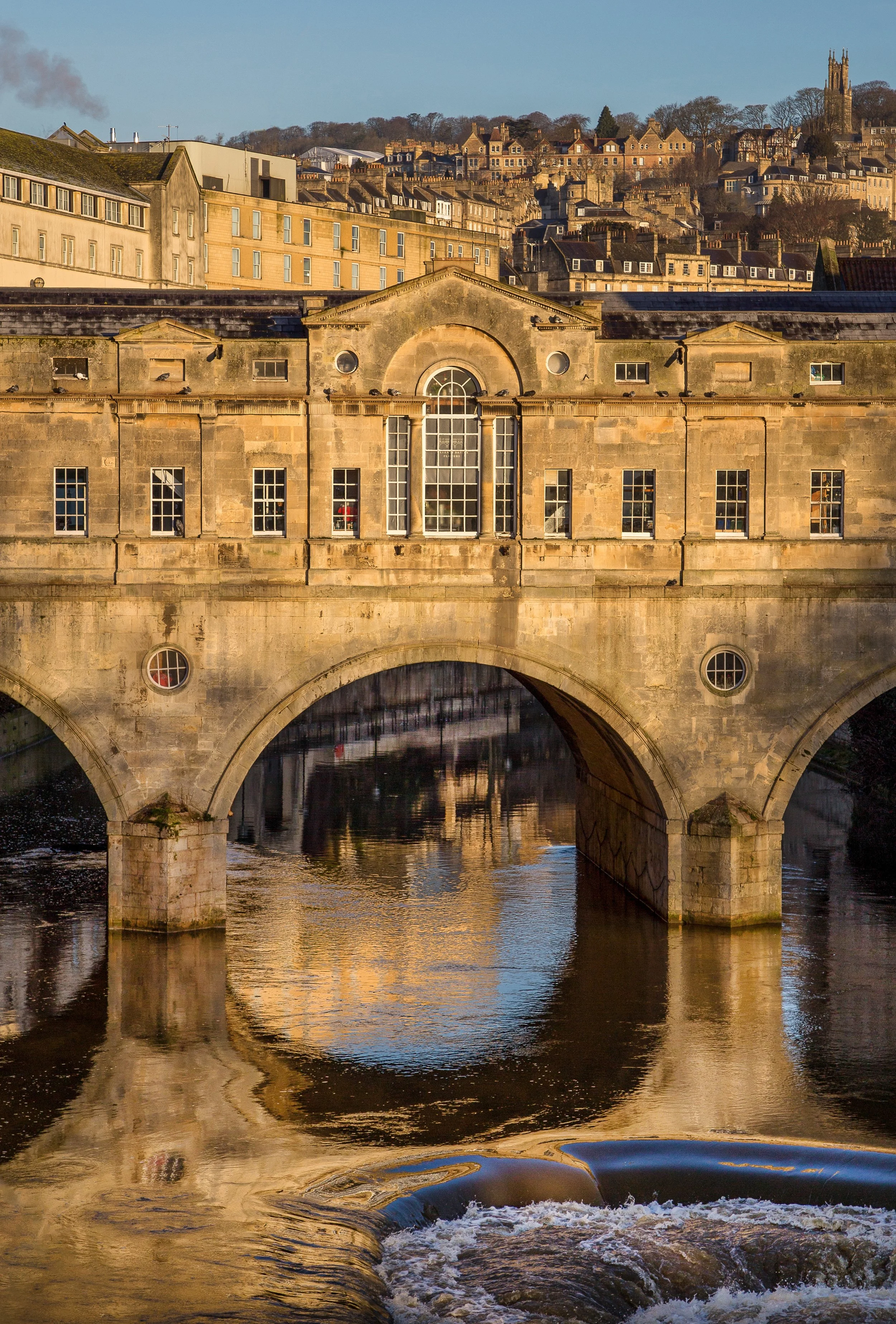Historic stone bridge over a river with reflections, surrounded by old buildings and a hillside with houses in the background, under a clear blue sky.
