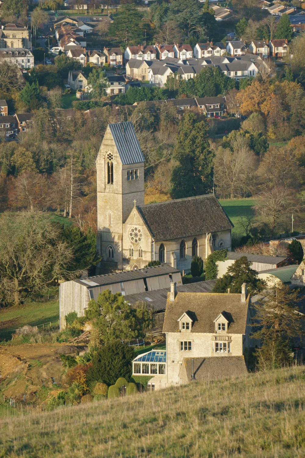 Aerial view of a small church with a tall steeple and circular stained glass window, surrounded by residential houses and trees in a hilly, suburban area.