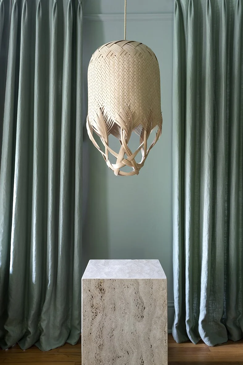 A woven pendant light hanging above a stone pedestal, with green curtains in the background. Bryony Richardson Interior Design, Jersey.