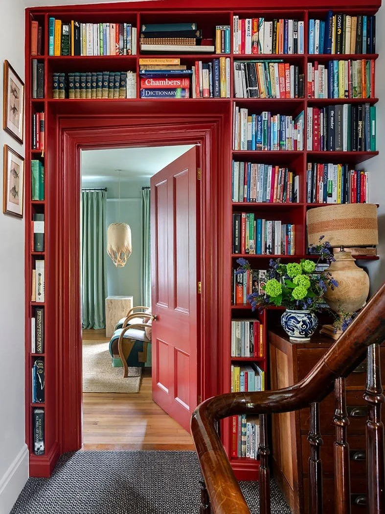 Bookshelves filled with colourful books frame a red-painted doorway, leading into a room with green curtains, a wooden chair, and a hanging lamp. Bryony Richardson Interior Design, Jersey.
