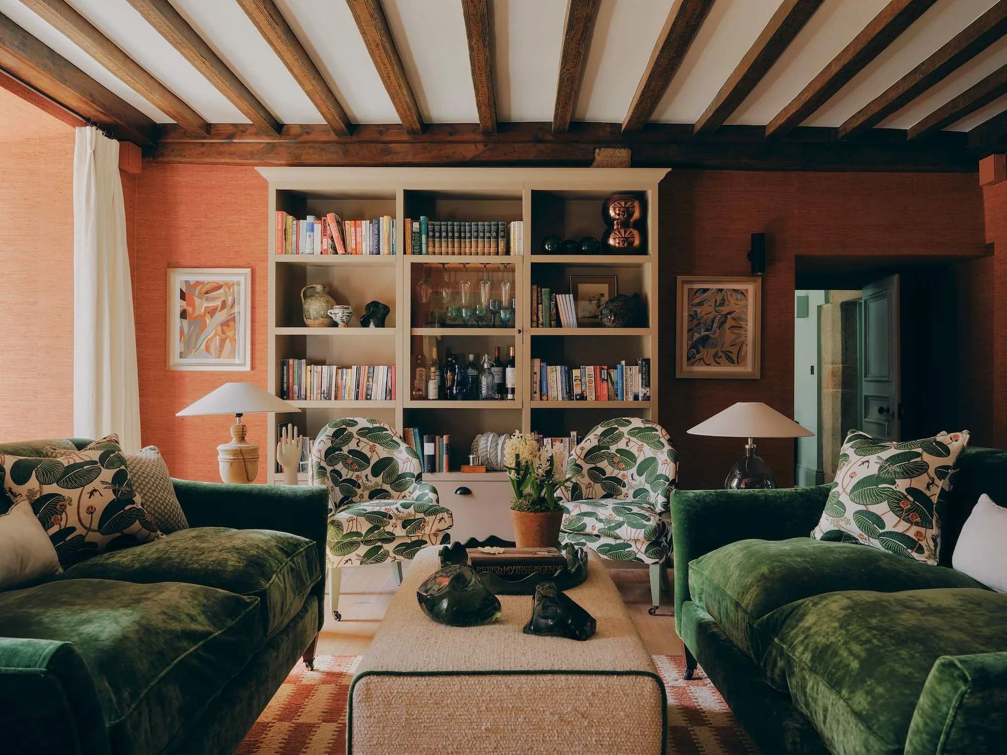 Living room with green velvet sofas, patterned armchairs, a coffee table with a plant, a white bookshelf with books and decorative items, framed art on the walls, and wooden ceiling beams. Bryony Richardson Interior Design, Jersey.