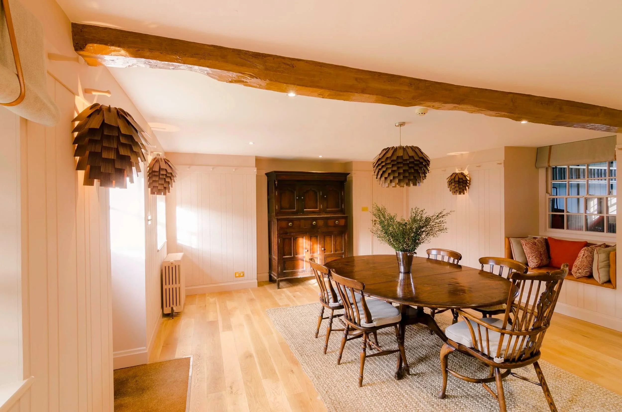 "Farmhouse breakfast room with an oval dining table, exposed beam and pinecone pendant lights in a Jersey granite farmhouse, designed by Bryony Richardson"