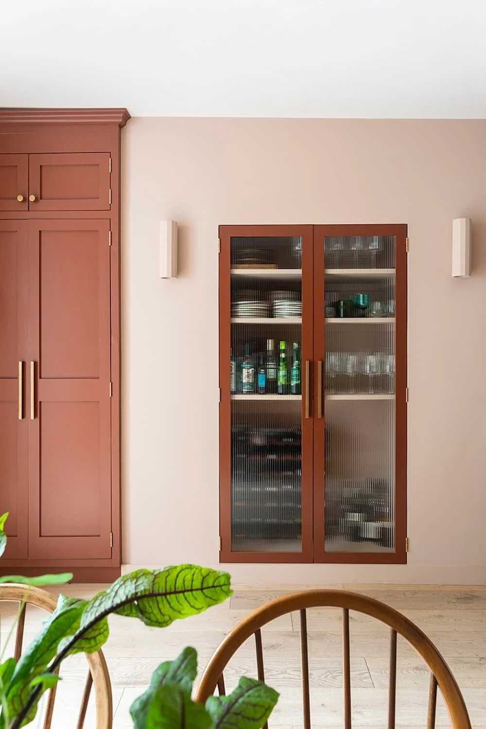 Kitchen cabinet with glass doors holding bottles and dishes, set against a pink kitchen wall, with a plant in the foreground. Bryony Richardson Interior Design, Jersey.