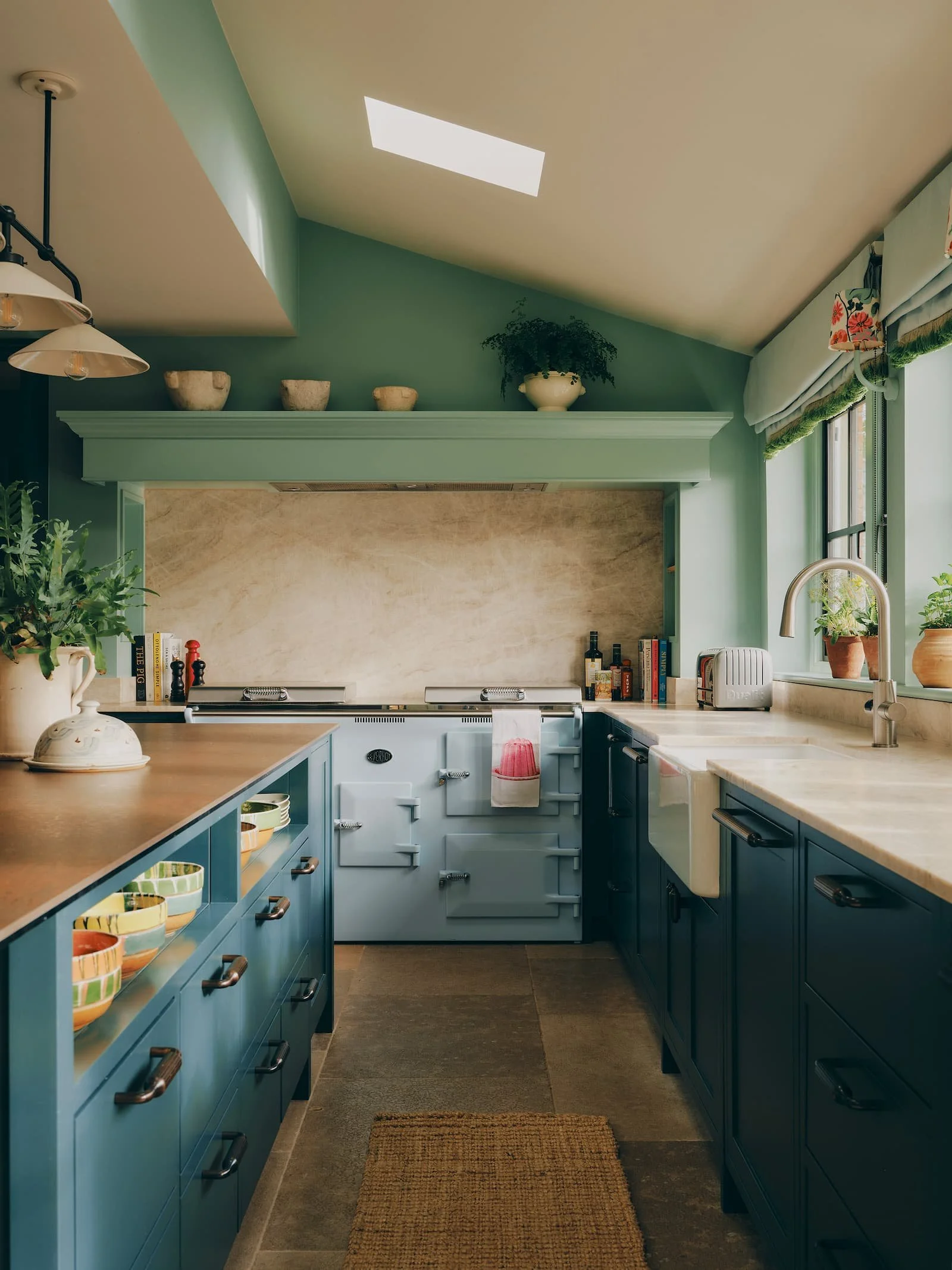 A cosy kitchen with blue cabinets, green walls, a beige countertop, and decorative bowls. There are potted plants on the windowsill, and a skylight provides natural light. Bryony Richardson Interior Design, Jersey.