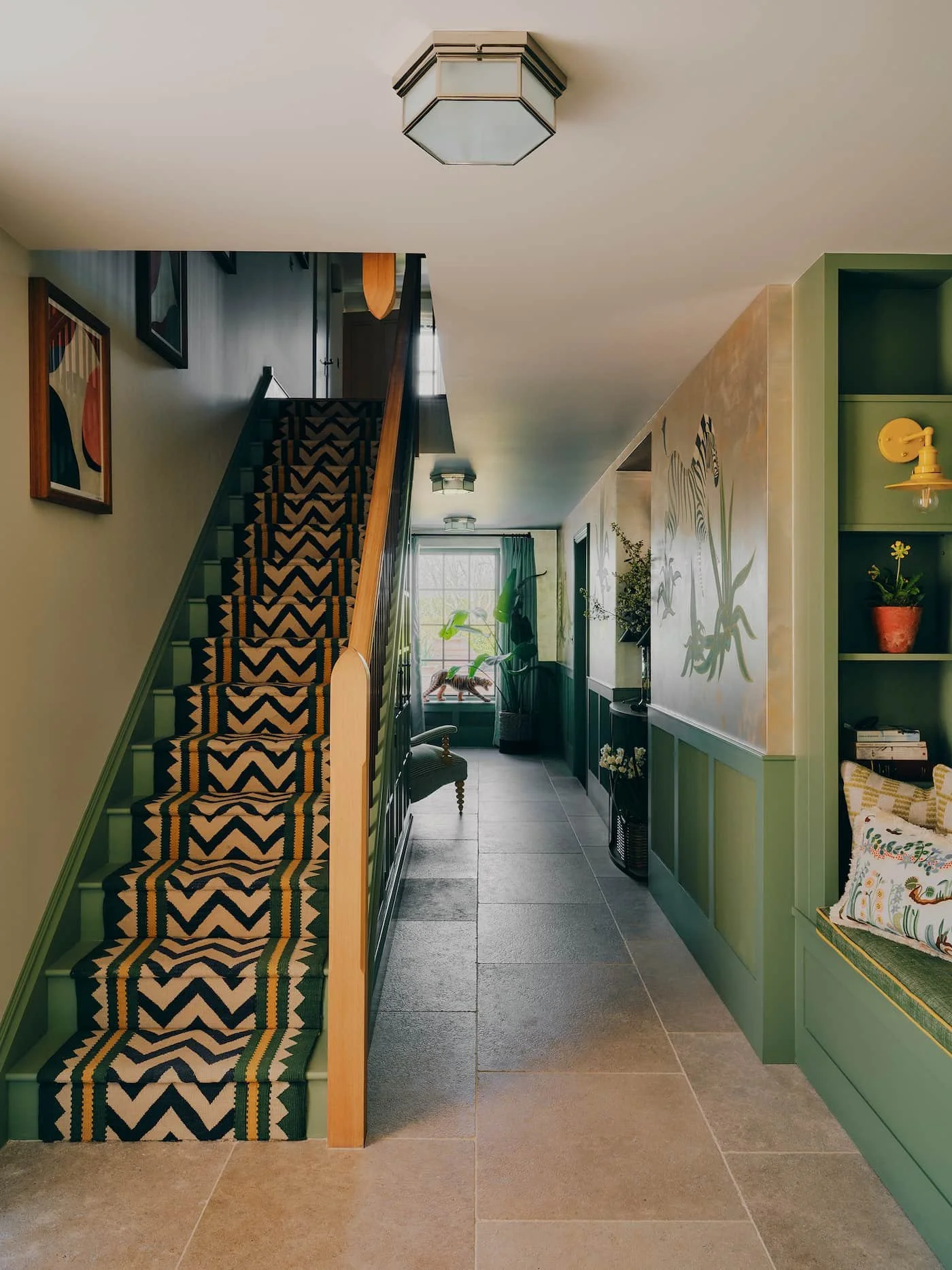 Interior view of a hallway with a staircase, framed artwork on the wall, potted plants, and a window at the end, with a green colour scheme and decorative lighting fixtures. Bryony Richardson Interior Design, Jersey.