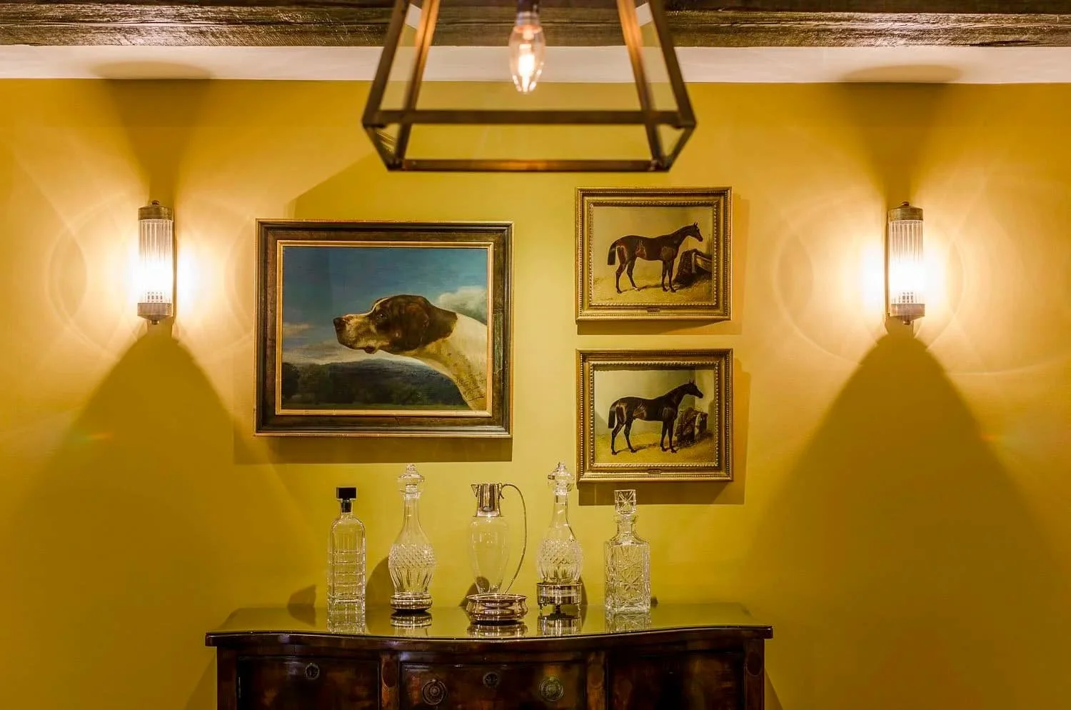 "Dining room wall with sporting dog and horse paintings, brass sconces and a sideboard of cut-glass decanters in a Jersey granite farmhouse, designed by Bryony Richardson"