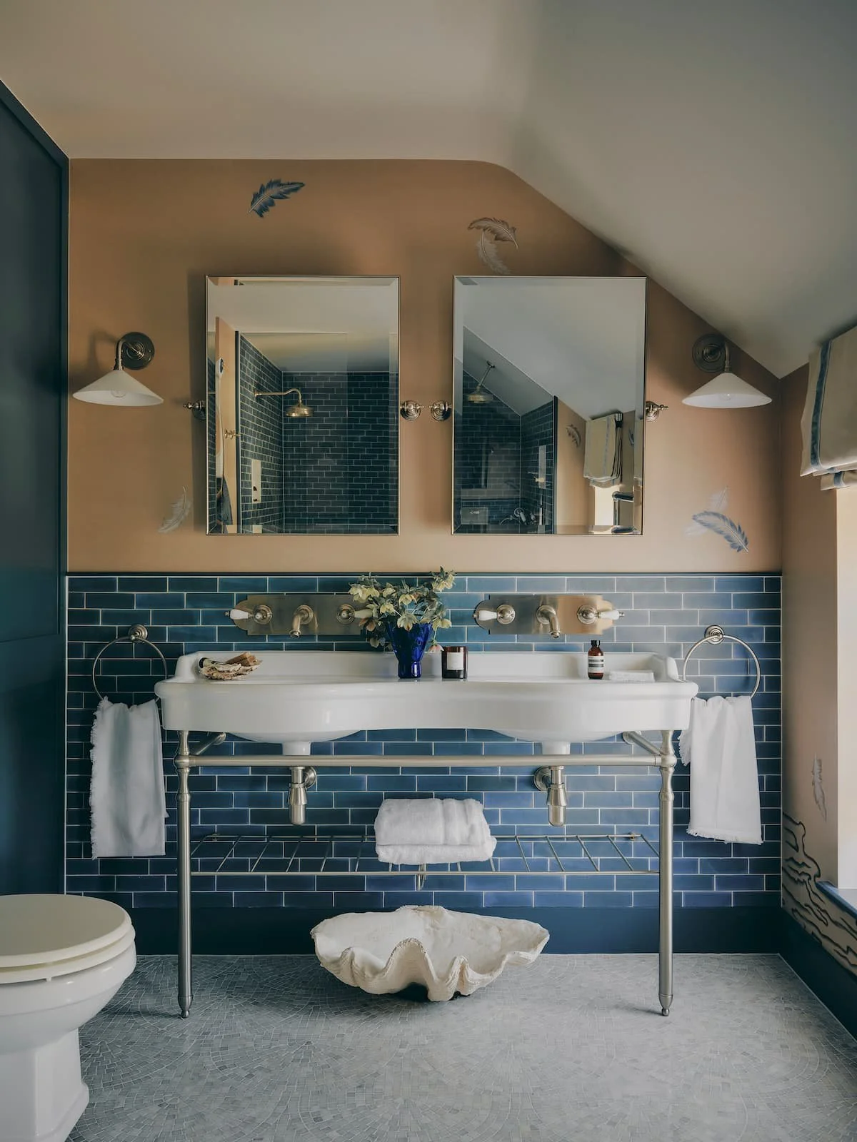 A bathroom with a double sink, blue subway tile backsplash, and two mirrors. There are wall-mounted lights and decorative feathers on the walls. The vanity has a blue vase with flowers, and towels hang from rings. A shell-like container is beneath the sink, and a shower with dark tile is visible in the mirrors.