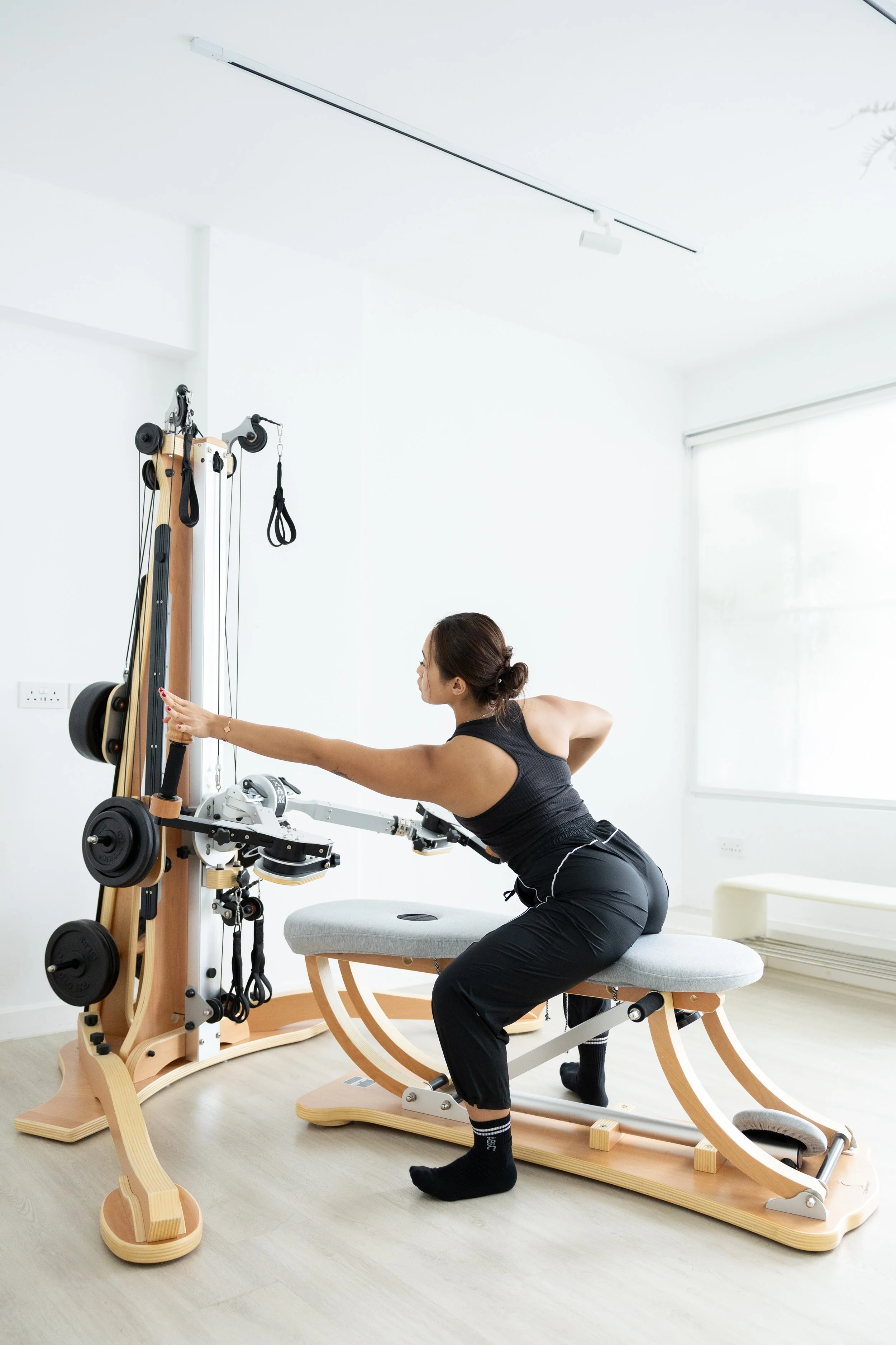 Woman exercising on Pilates reformer machine in a bright, modern room with minimal decor and large window.
