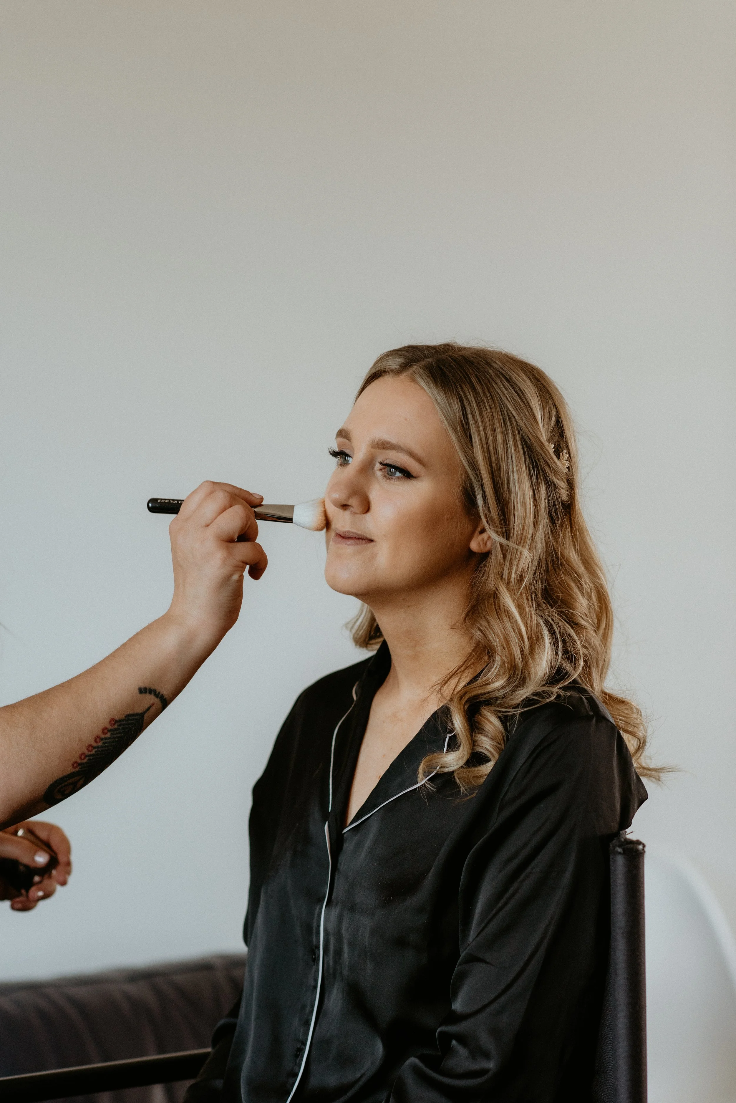 A woman with wavy blonde hair getting her makeup applied with a brush by a makeup artist.