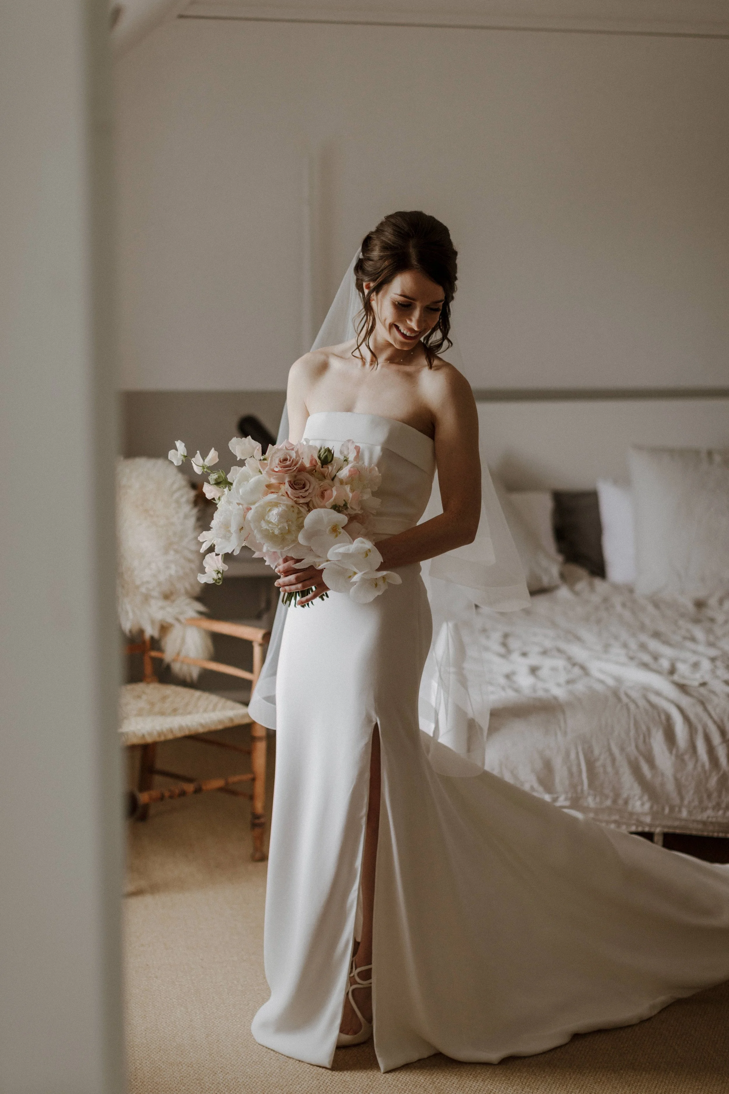 A bride in a strapless white wedding dress holding a bouquet of pink roses and white flowers, standing in a bedroom with a bed and a chair in the background.