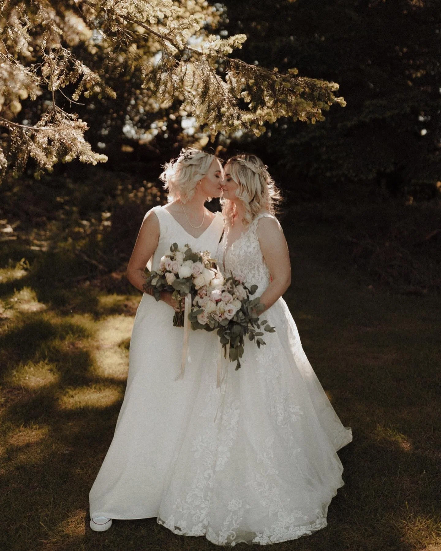 Two women in wedding dresses sharing a tender moment outdoors surrounded by trees, holding a bouquet of flowers.