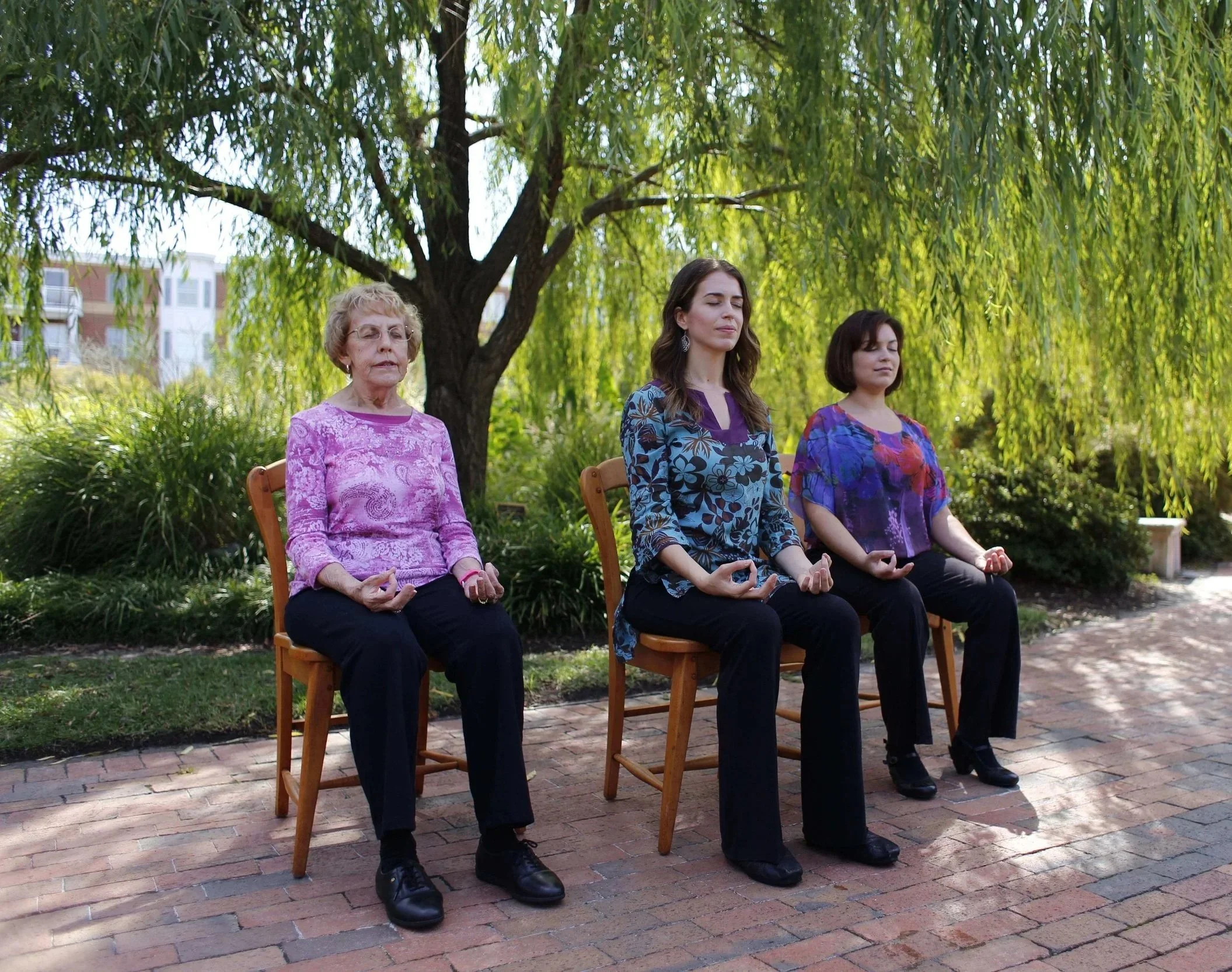 Ann sitting on a wooden chair outdoors with trees in the background, eyes closed and hands in a mudra pose, alongside an elderly woman on the left and another woman on the right doing the same.