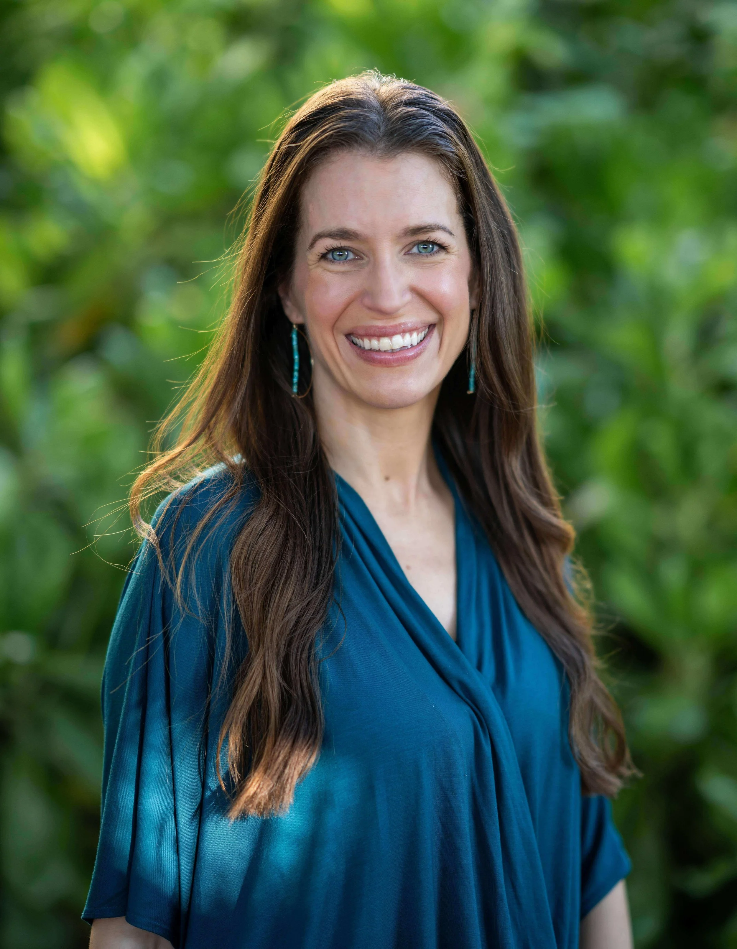 Ann smiling at the camera, wearing a  teal shirt, hair down, with a nature backdrop.