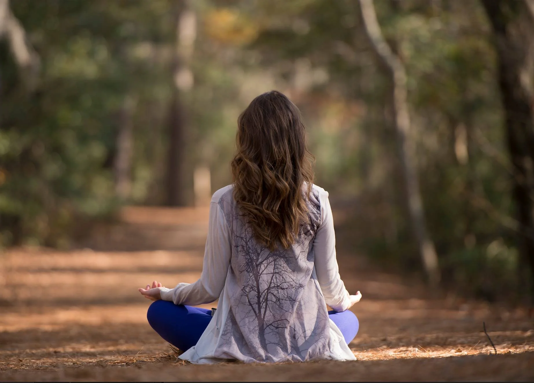 Ann sitting in lotus position with hands on her knees, outdoors in nature, facing away from the camera.