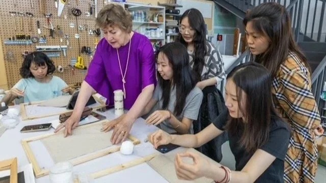 A group of five women, including a woman with curly hair wearing a purple top, are working on a craft project together at a table in a workshop or classroom. They are surrounded by workshop tools and materials.