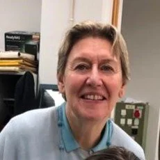 Smiling woman in a blue shirt, sitting in an office with shelves and equipment in the background.