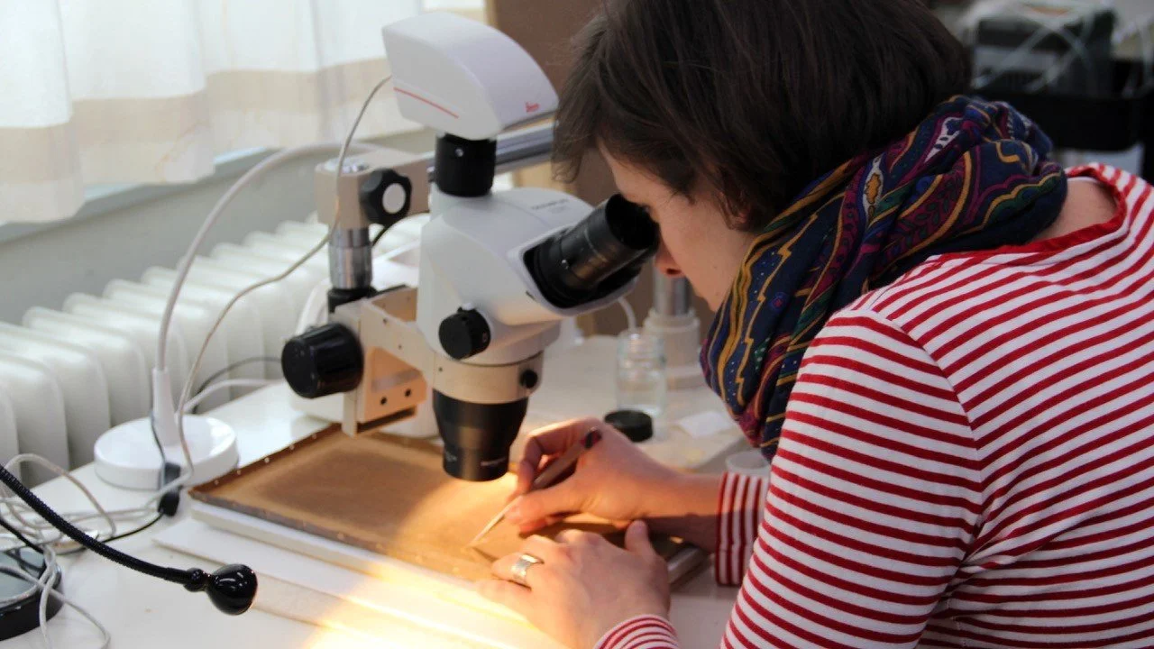 A woman in a red and white striped shirt and a colorful scarf is looking through a microscope while working with a small tool on a piece of wood or material.