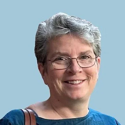 A woman with short gray hair, glasses, and a blue shirt smiling against a light blue background.