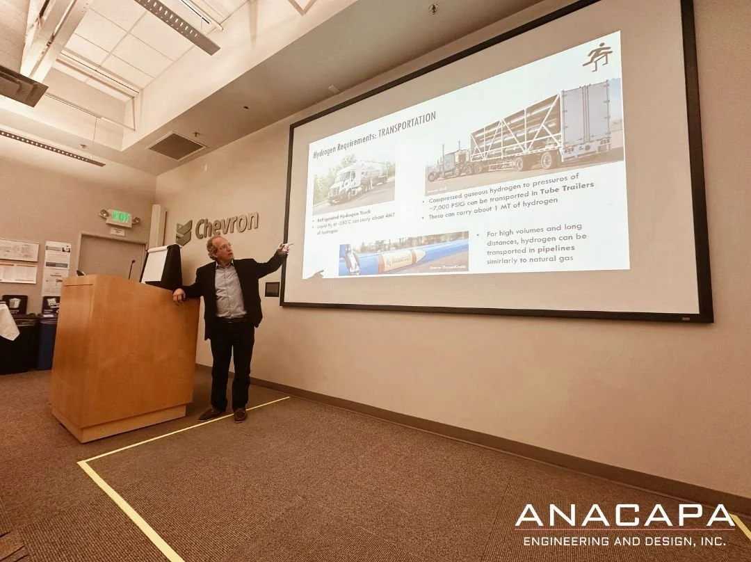  Stuart M. Heisler, PE. stands at a podium giving a presentation in a room. An image of a large truck and text on hydrogen transportation is displayed on the screen. 