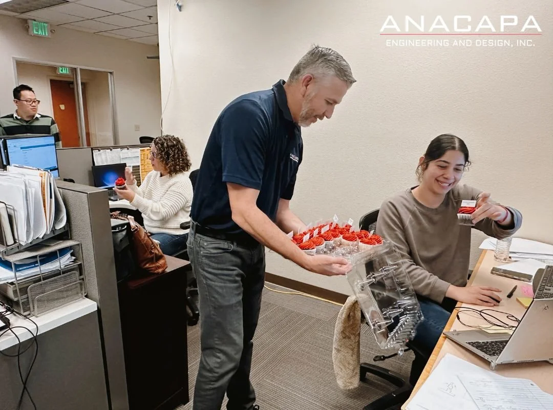  Office scene with a man handing out tasty treats to a smiling woman at her desk in celebration of Engineers Week. Other employees are working in the background, and the company logo "ANACAPA Engineering and Design, Inc." is visible in the top corner