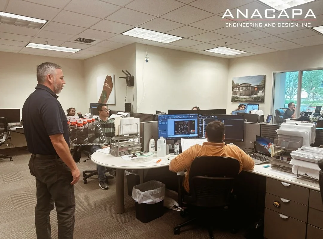  A group of people working at their desks with computers at Anacapa’s office. A man stands near the desks, and the office features various design tools and wall-mounted artworks. "ANACAPA" is visible in the top right corner, celebrating Engineers' We