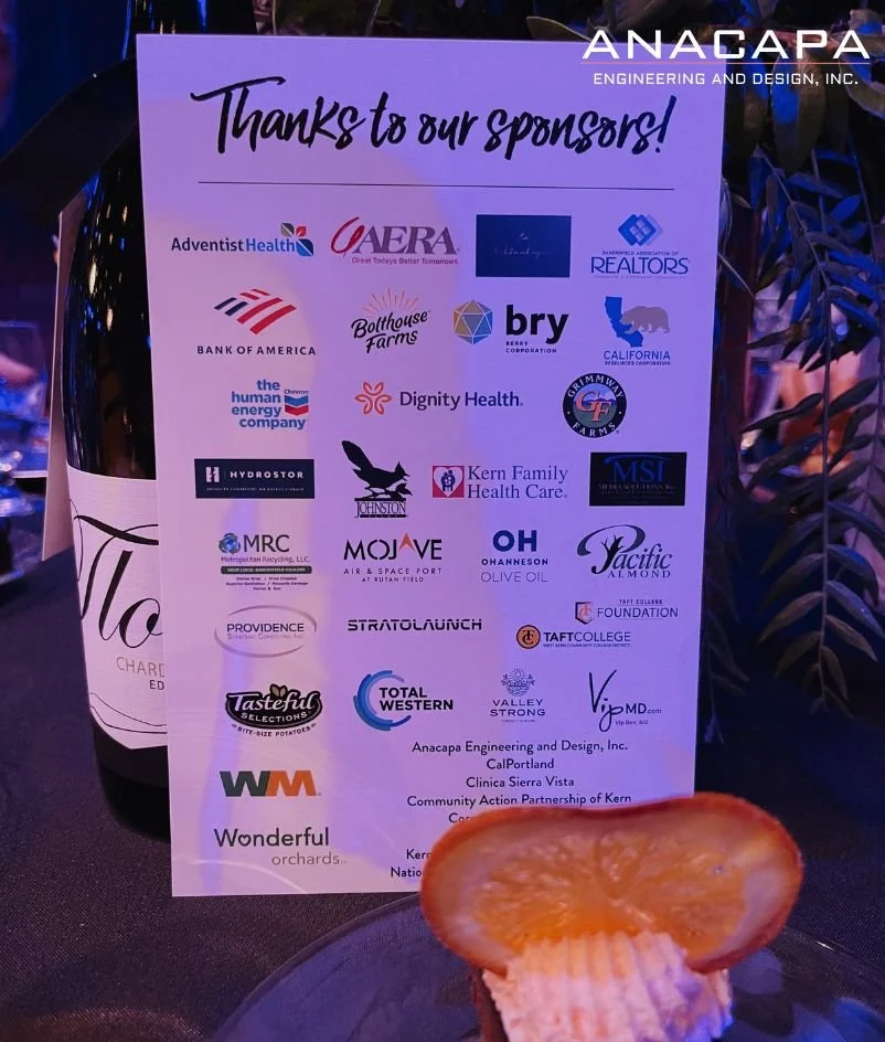  A sponsorship board displaying various company logos, a bottle of white wine, and a plated slice of food in the foreground. The board is titled "Thanks to our sponsors!" as part of the State of the County event hosted by Anacapa. 