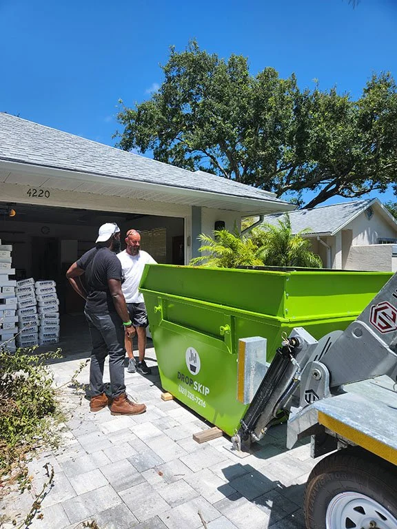 Two men are standing outside a house near a green trash container on a trailer, with one man wearing a white cap and a black shirt, and a large tree and clear blue sky in the background.