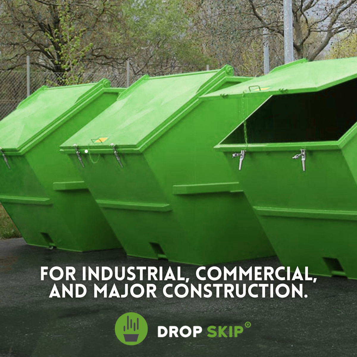 Green industrial dumpsters lined up outdoors on a paved surface with trees in the background.