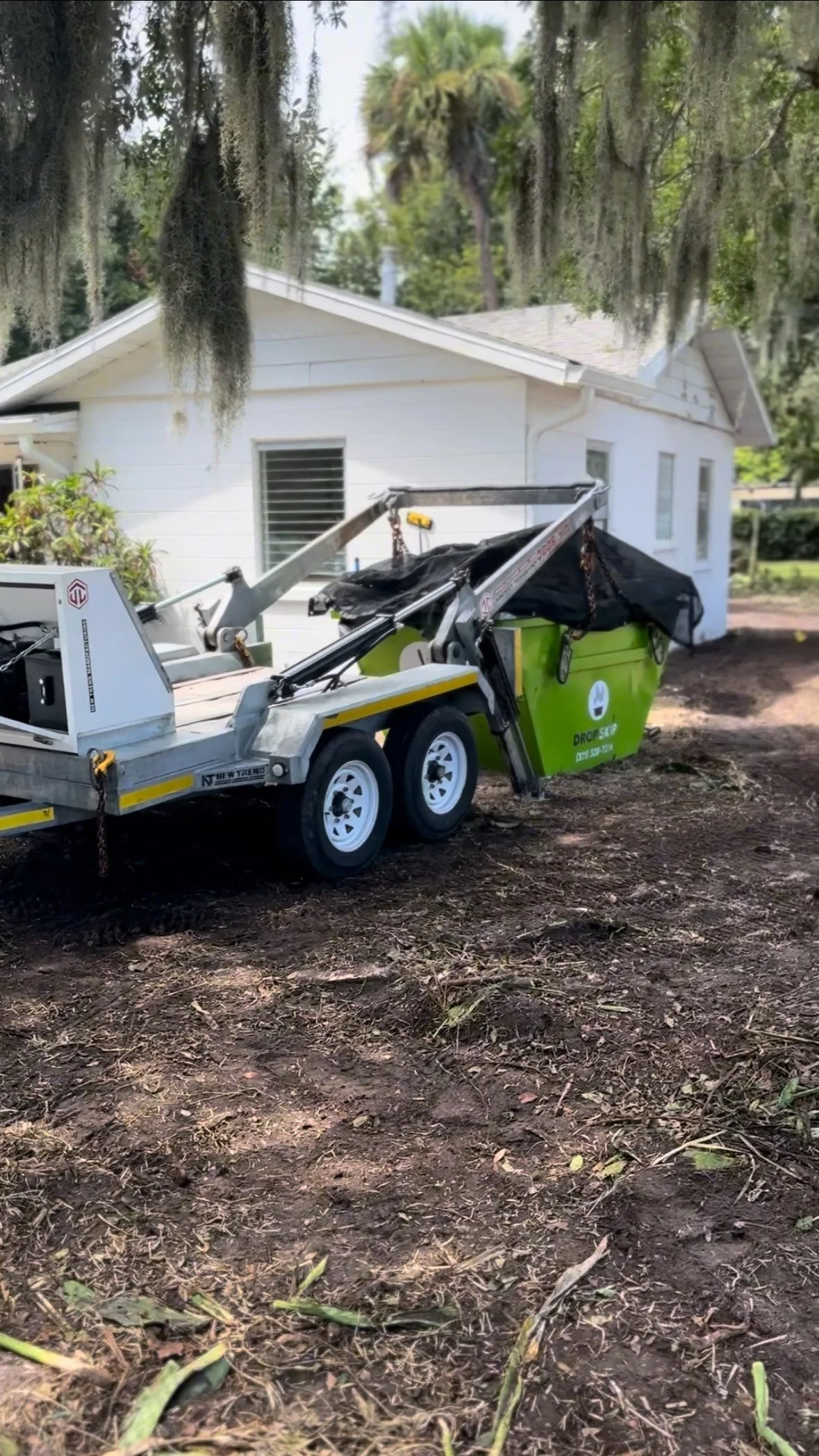 A green and gray drop off trash container attached to a trailer, parked outside a white house with trees overhead.